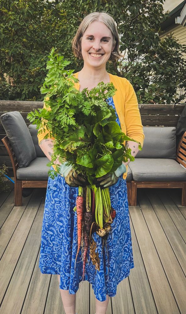 Photo of Jen standing on a deck wearing a blue dress, yellow cardigan and holding a freshly picked bunch of garden carrots and beets. 
