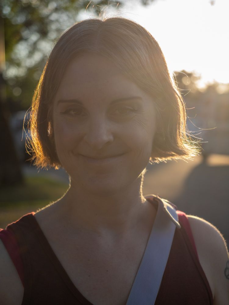 Photo of smiling Jen on a tree lined street with sunbeams.