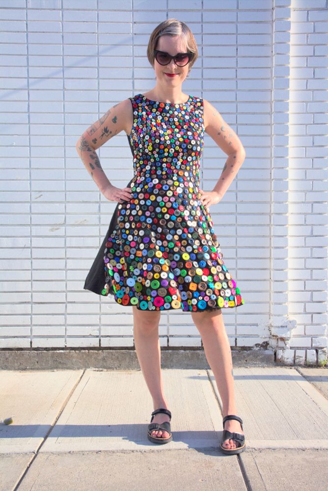Photo of Jen from August 2020 standing outside against a white brick wall wearing a black dress covered in 1200 colourful buttons. 