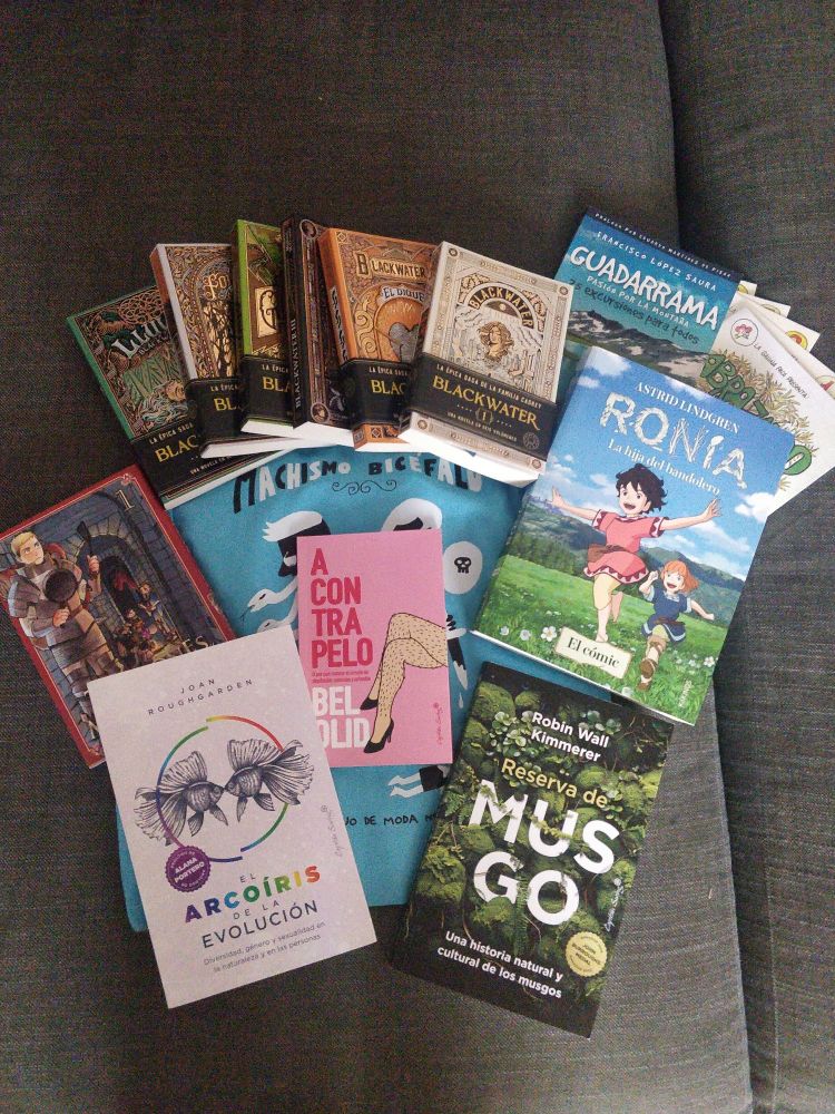 Montón de libros sobre una totebag azul turquesa: 
los seis tomos de Blackwater, guia de excursiones por la sierra de Guadarrama y laminas con dibujos de fauna y flora, primer tomo de Tragones y Mazmorras, A Contrapelo (Bel Olid), el arcoiris de la evolución (Joan Roughgarden) y Reserva de Musgo (Robin Wall Kimmerer).