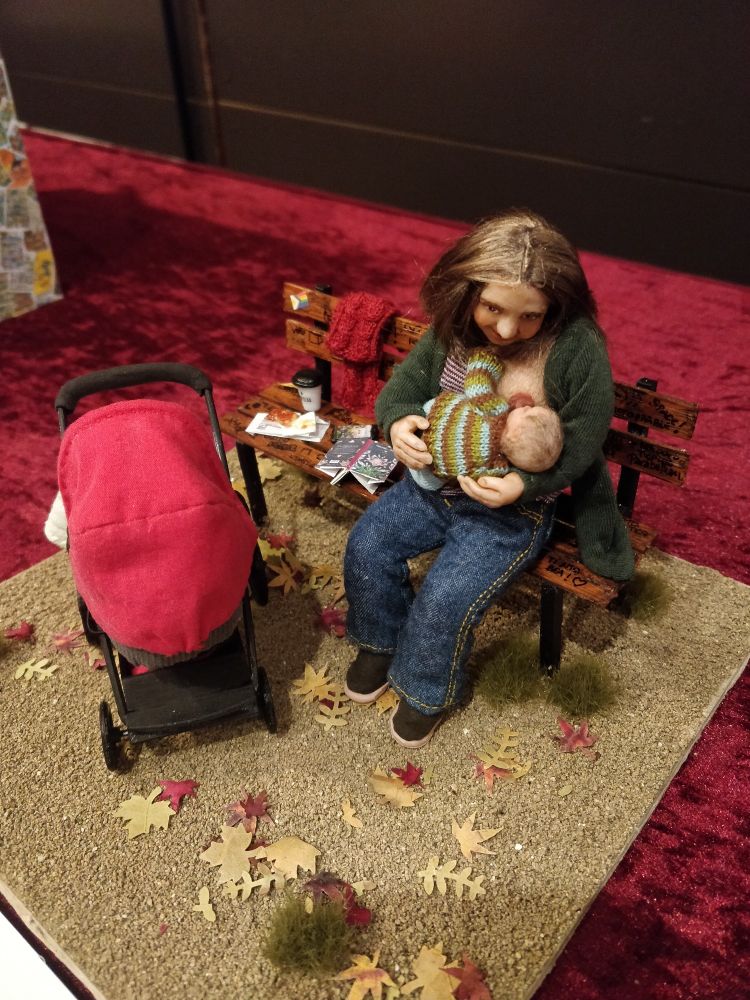 Escena 1:12 exterior, un banco del parque y el suelo lleno de hojas secas. Una mujer da el pecho a un bebé, junto a un carro con capazo y la capota roja. A su lado en el banco un libro abierto bocabajo, un dulce y un café para llevar, y colgada en el respaldo una bufanda roja