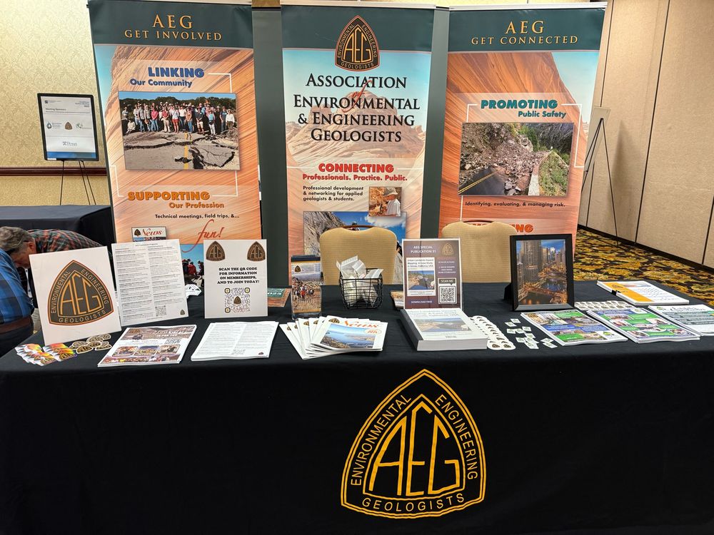Association of Environmental and Engineering Geologists exhibit booth with flyers, pop-up signs, stickers on a table with black tablecloth and yellow AEG logo