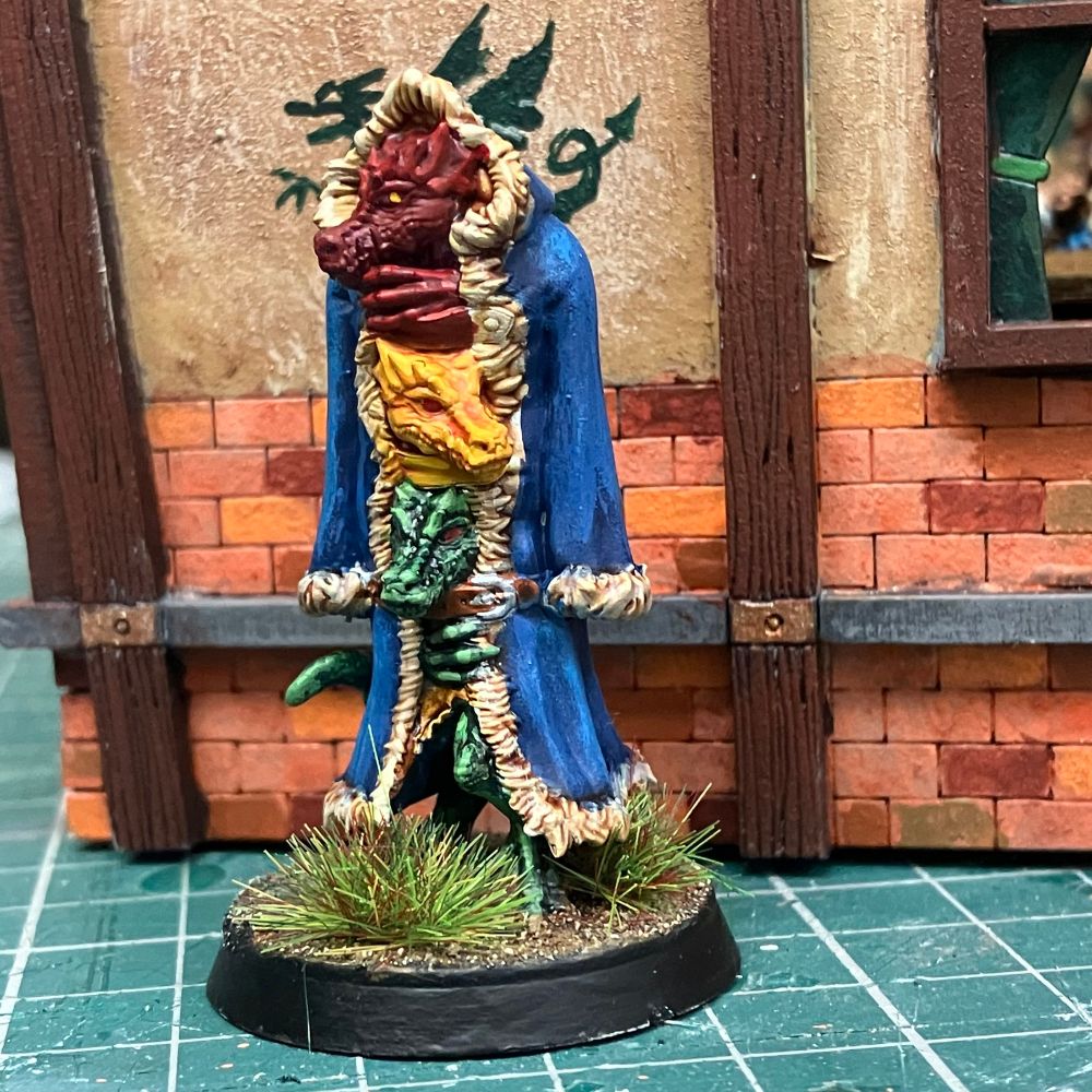 Three kobolds are standing on each others shoulders, partially concealed by a hooded blue coat with white fur trim. The top kobold is red, the middle yellow, the bottom green. A green tail is also poking out of the back of the coat. 