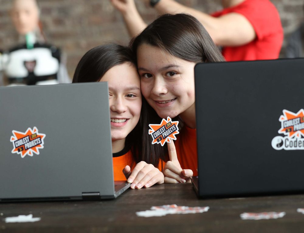 Two young people sitting behind laptop screens holding up a "Coolest Projects" sticker at a previous in-person Coolest Projects event.