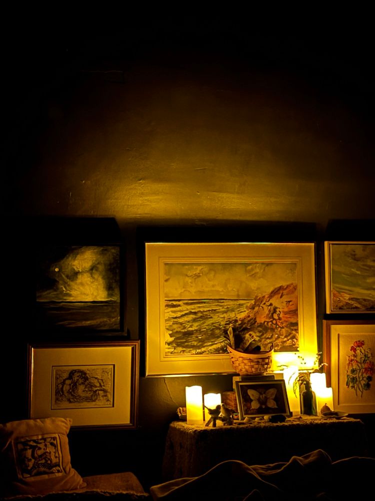 A small table covered with vintage crochet tablecloth and small personal mementos as well as remote control candles. On the wall behind the table are several paintings. 