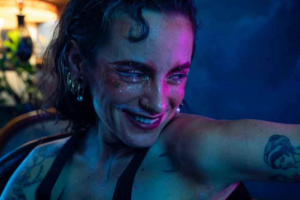 Une personne souriante sous éclairage bleu et rose, avec du maquillage brillant avec paillettes et étoiles autour des yeux. Tatouages visibles : lune sur l’aisselle et visage féminin sur le bras. Boucles d’oreilles perlées et anneau nasal ajoutent une touche élégante et alternative.