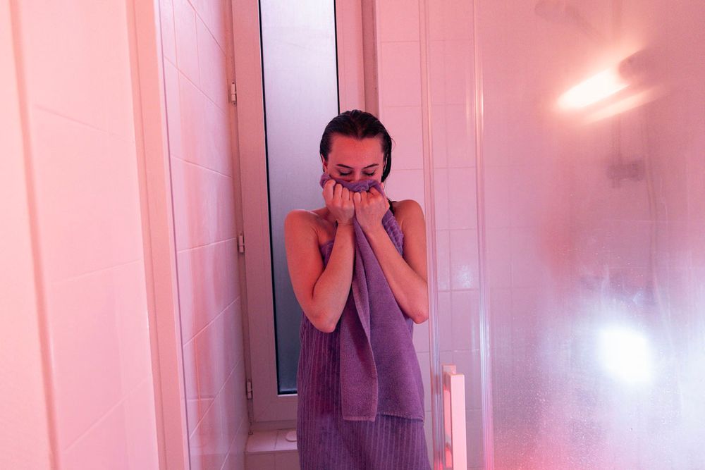 Femme sortant de la douche, emmitouflée dans une serviette violette, le tout dans une lumière aux tonalités roses.