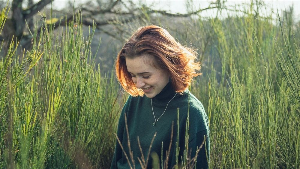 Portrait de femme souriante dans un champs entouré de plantes vertes. Le soleil fait briller la rousseur des cheveux.