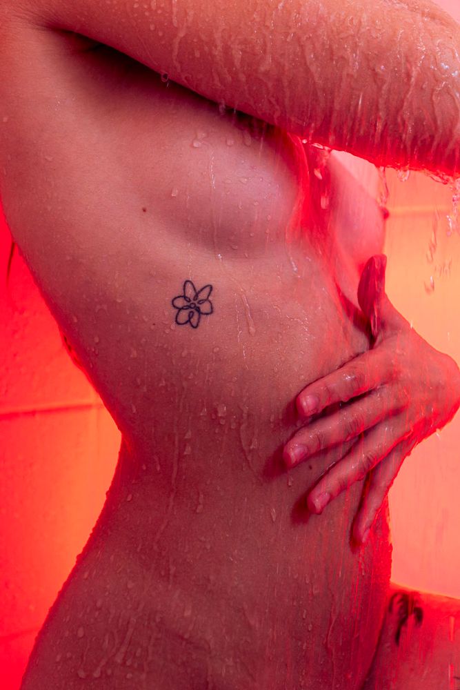 A close-up of a person's torso under running water, showing a simple flower tattoo on their ribcage, with an orange-red light illuminating the scene.