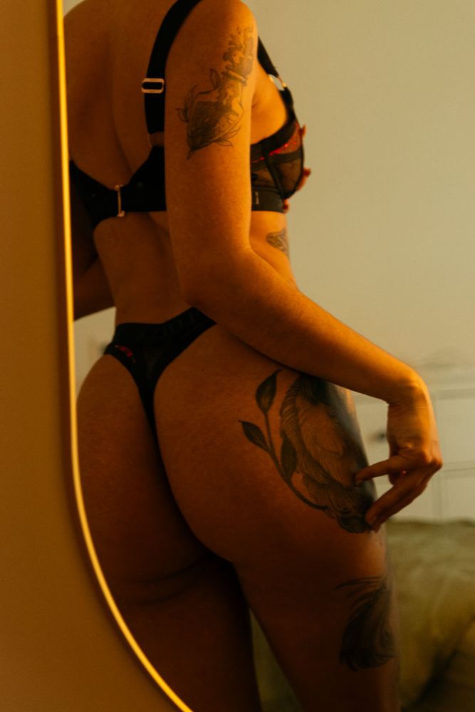 A person wearing black lingerie with visible tattoos poses in front of a mirror in a dimly lit room.