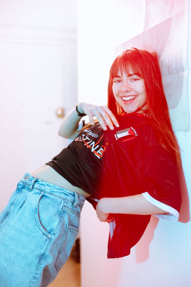 Une jeune femme rit en se penchant en arrière contre un mur, baignée dans une lumière rouge et bleue. Elle porte un t-shirt ample et un jean clair, créant un contraste décontracté avec l’éclairage vif. L’image respire la spontanéité et la bonne humeur.