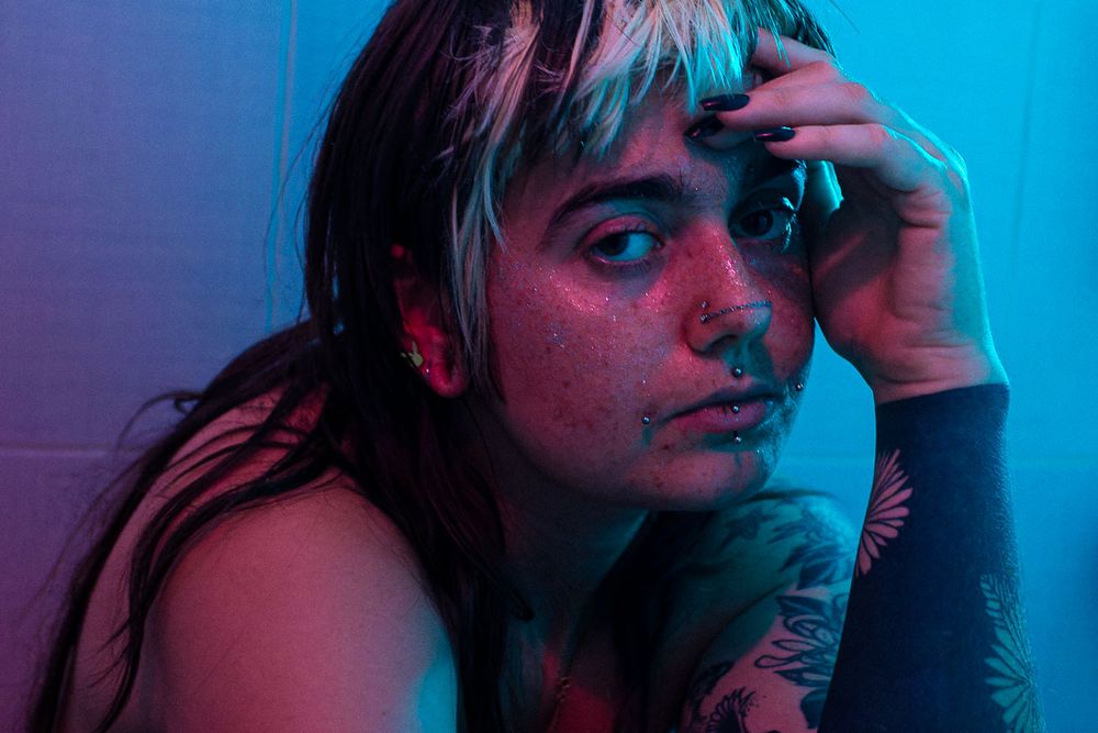 Portrait de femme recroquevillée sous la douche, éclairée par une lumière rose (et bleue en arrière plan). Elle porte des piercings sur le visage et un grand tatouage floral sur le bras.