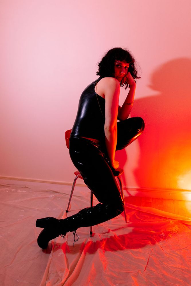 A person in a black, shiny outfit poses on a red chair in a room with red and yellow lighting and a plastic-covered floor.