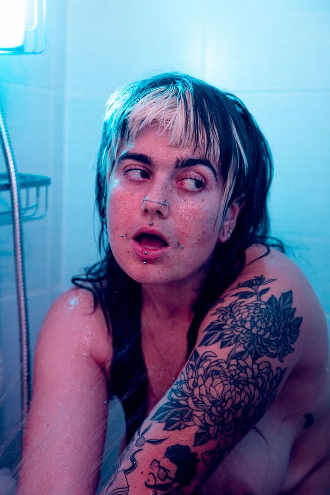 Portrait de femme recroquevillée sous la douche, éclairée par une lumière rose (et bleue en arrière plan). Elle porte des piercings sur le visage et un grand tatouage floral sur le bras. L’expression est intense, presque rêveuse.