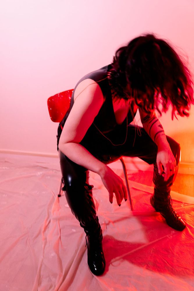 Un portrait dynamique sous éclairage rouge, où la personne en cuir noir et bottes hautes se penche en avant, cheveux masquant le visage. L’attitude et la mise en scène dégagent une énergie brute et théâtrale.