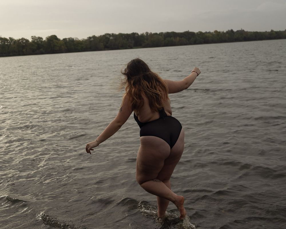 Silhouette féminine de dos en body noir, qui s'aventure dans un lac naturel. La lumière grise et orageuse contraste avec la légèreté du moment.