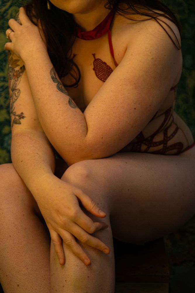 Plan rapproché d'une personne assise, partiellement vêtue de lingerie en corde rouge, dans une pose intime et artistique. La lumière est douce et chaleureuse, mettant en valeur les détails du corps.