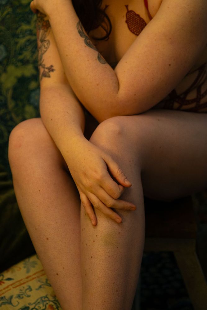 Plan rapproché d'une personne assise, partiellement vêtue de lingerie en corde rouge, dans une pose intime et artistique. La lumière est douce et chaleureuse, mettant en valeur les détails du corps. La main effleure le genou.