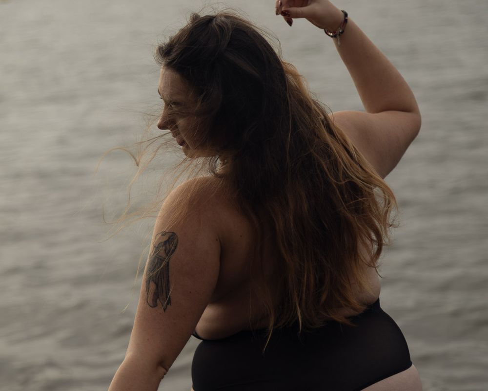 Silhouette féminine de dos en body noir, qui s'aventure dans un lac naturel. La lumière grise et orageuse contraste avec la légèreté du moment.
