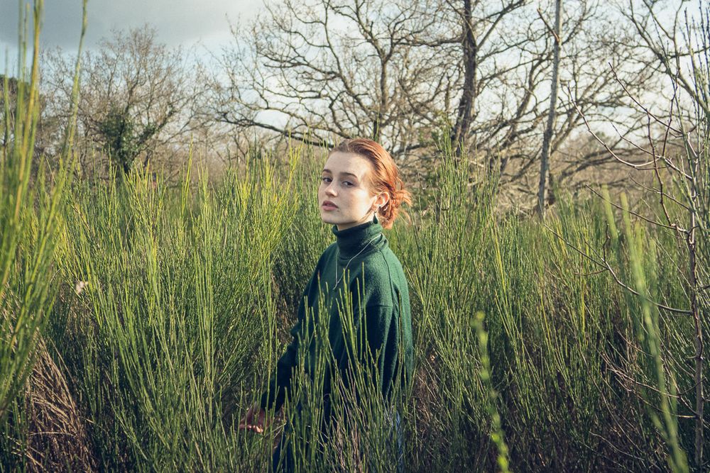 Portrait féminin dans un champs entouré de plantes vertes. Le soleil fait briller la rousseur des cheveux.
