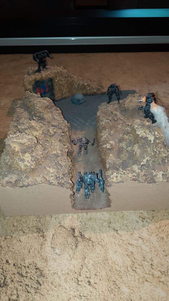 A diorama of a canyon leading to a gate built in the rocks, with a turret near the gate. Three Battlemechs on top of the canyon walls attacking two Mechs walking through the canyon.
