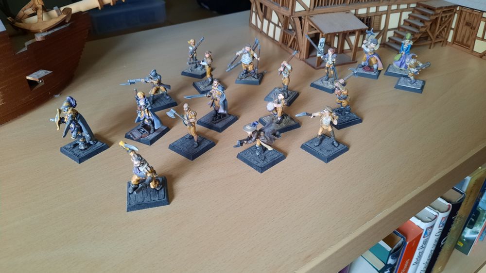 Some Freebooters Fate Imperial Armada Miniatures standing on the top of a wooden shelf. Behind them a part of a Tavern Model set on the right side and a part of a ship set on the left side.