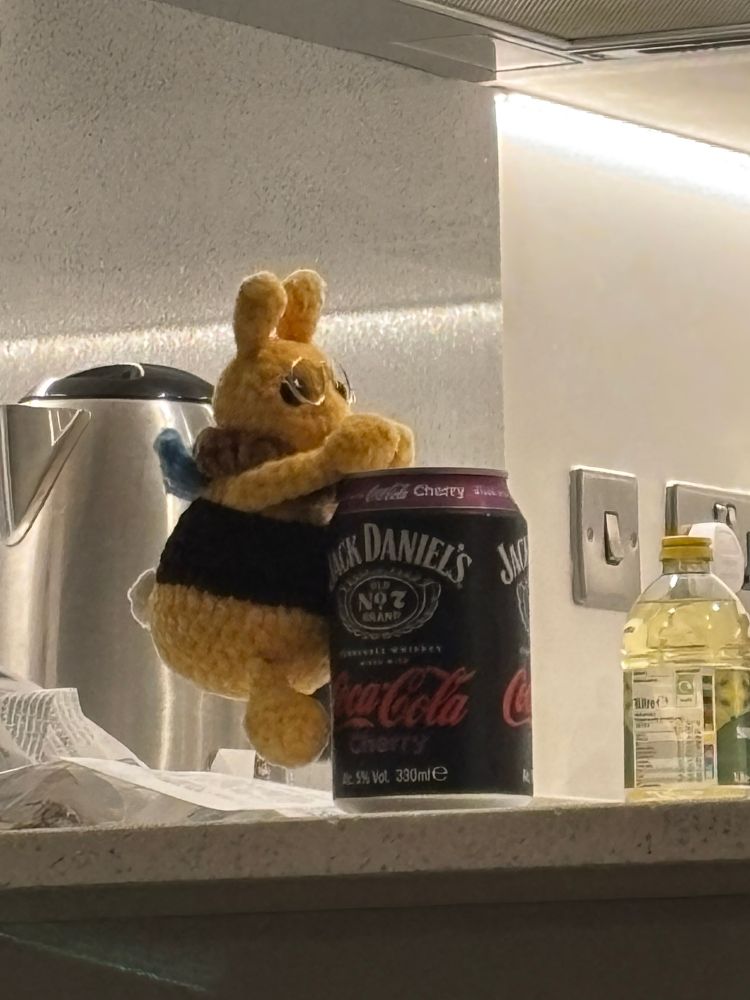 zoomed on crocheted bilbee is sitting on the kitchen counter with his arms around a can of whiskey and coke