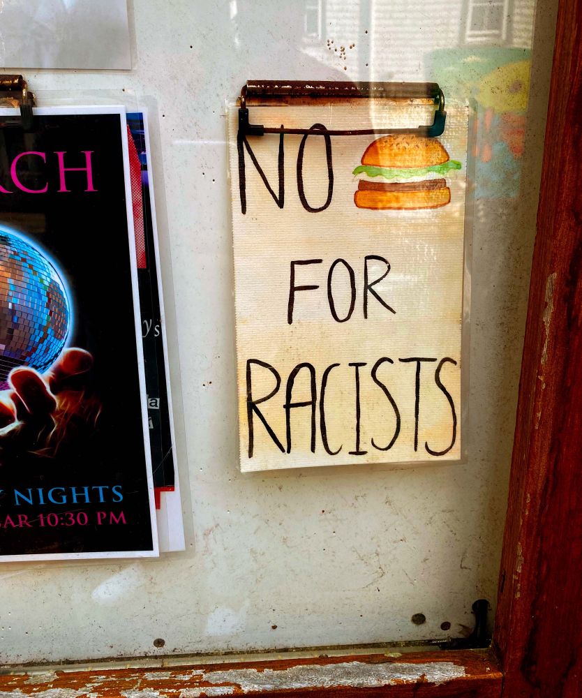 A handmade sign that has “NO BURGER FOR RACISTS” written on it. A hamburger on a sesame seed bun with lettuce and onion has been drawn instead of the word burger. The sign is encased in glass with a worn wooden frame along the outer edge, and hangs alongside other marketing materials. The reflection of another building along the downtown strip of Provincetown can be seen in the glass.