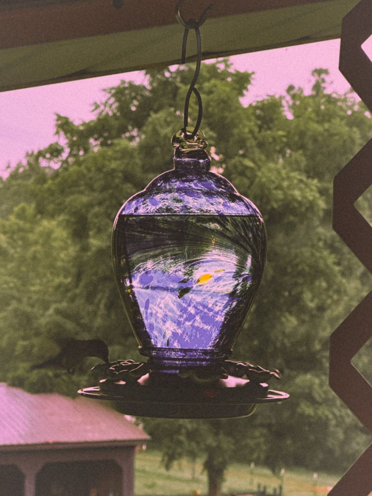The blue stained glass hummingbird has a hummingbird flying on the left sipping from the feeder. Part of a barn with a large tree can be seen in the background.