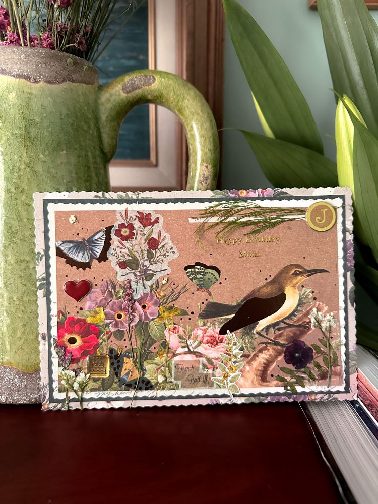 A view of just the handmade card which is oriented horizontally and features a blue butterfly in the upper left, a variety of pressed flowers along the bottom of a flower printed piece of cardstock, a bird resting on a branch in the lower right, a green butterfly in the middle, and wispy greenery in the top right with a round gold and brown “J” sticker and a pink polka dot line going partially across the top of the card. The card has a very strong vintage floral theme, and there are small features that represent my mother throughout. 
