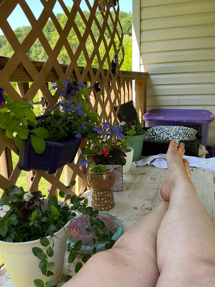 A woman has her legs crossed on top of a wood garden bench outside, and various flower arrangements hang on a trellis and sit on top of the bench. A chair, bins for soil storage, and a lush green Mountain View can be seen