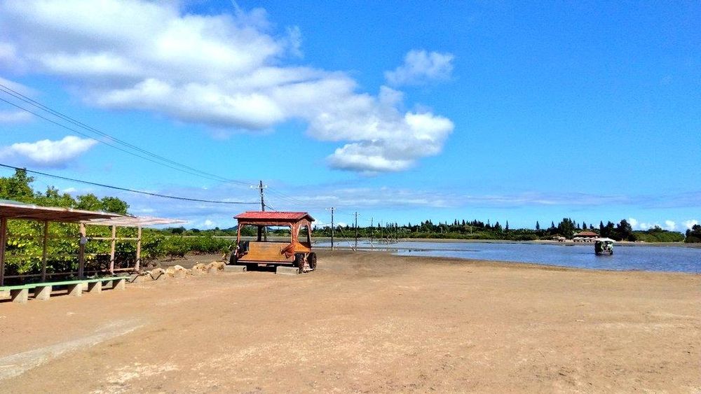 青空の下、川沿いの広場に木製の屋根付き荷車が停まっており、右奥には小さな船が水面に浮かんでいる静かな風景。