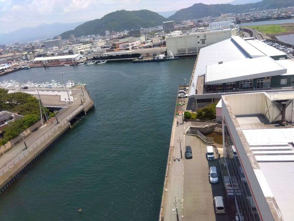 港に面した都市の風景。右側に倉庫や商業施設、左側に船が停泊する港と道路、奥に山々と市街地が広がっている。晴天で水面が青く輝いている。