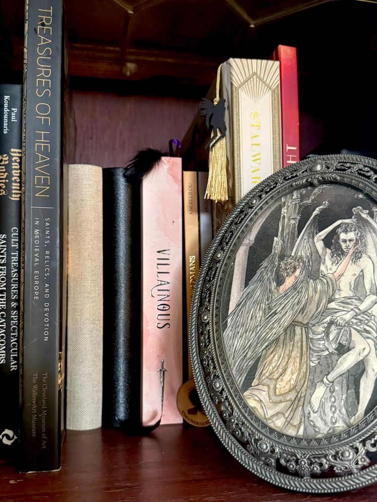 The all-fanwork, NG-free Good Omens portion of my bookcase: two books by FanBoundBooks, IneffablePenguin’s Villainous, Angellilou’s Ineffable Romans and Light Shades, dustandhalo’s Stalwart Sun, Rising Moon (with bonus bookmark also by IneffablePenguin), Illustory’s Let There Be Light, and a beautiful framed print by martina_a_duck.