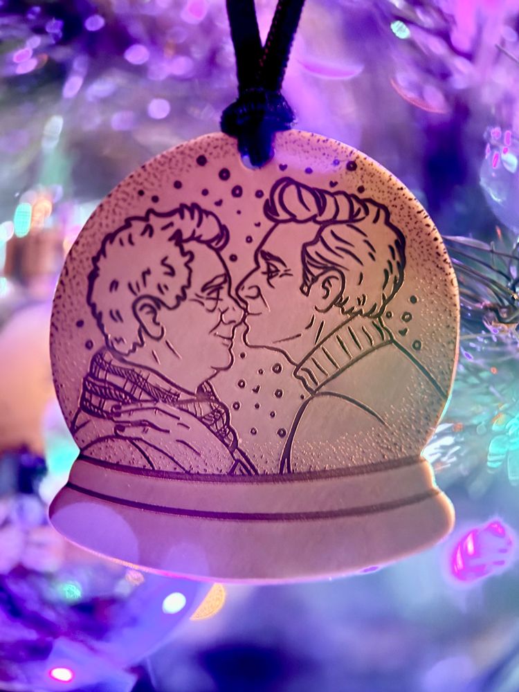 A copper ornament hanging on a tree surrounded by lights in shades of pink, purple, and blue. The ornament is shaped like a snowglobe, and engraved with art of Aziraphale and Crowley from the shoulders up, facing each other and surrounded by snowflakes. Crowley is wearing a turtleneck and leaning in with one hand on Aziraphale’s chest to kiss him on the tip of his nose. Aziraphale is wearing a tartan scarf and has his eyes closed with a gentle smile on his face.