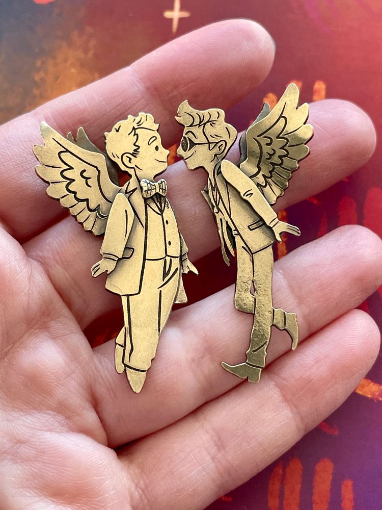A hand holding a pair of small layered brass brooches of Aziraphale and Crowley looking at each other.