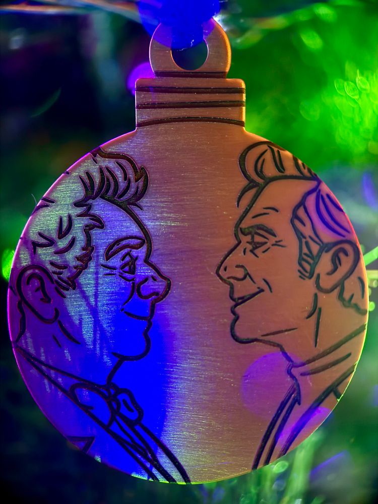A Christmas bauble-shaped brass ornament hanging in a tree, engraved with Aziraphale in profile on the left side and Crowley in profile on the right. They are looking at each other and smiling. The lights on the tree are casting a cool blue and green colors on Aziraphale and warm orange and pinks on Crowley.