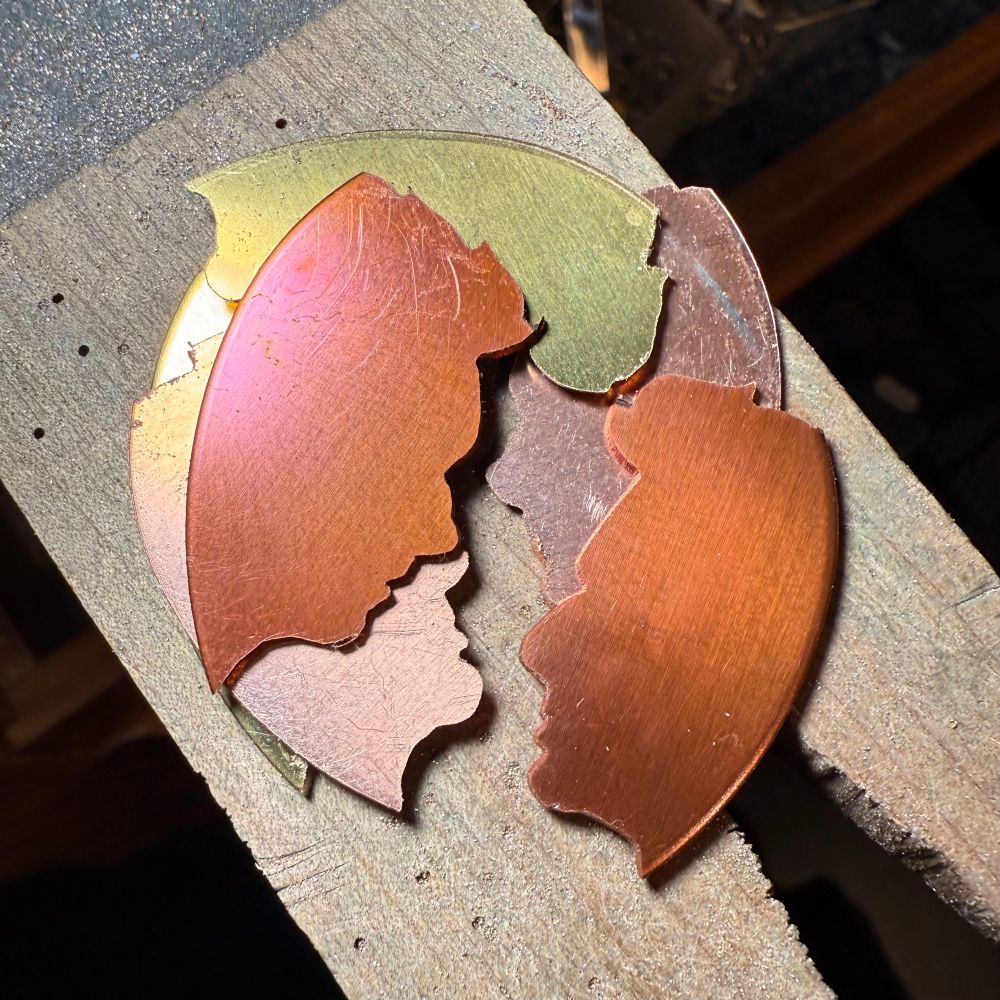 Silhouettes of Aziraphale and Crowley that have been sawed from copper and brass sitting on a wooden bench pin