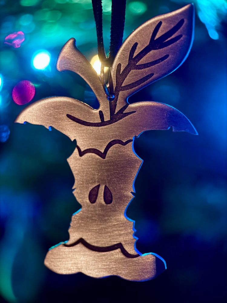 A brass ornament hanging on a tree surrounded by lights in shades of blues and greens. The ornament has silhouettes of Aziraphale and Crowley sawed out of either side, facing each other to create the illusion of an apple core. Some simple engraving suggests the stem, leaf, and seeds.