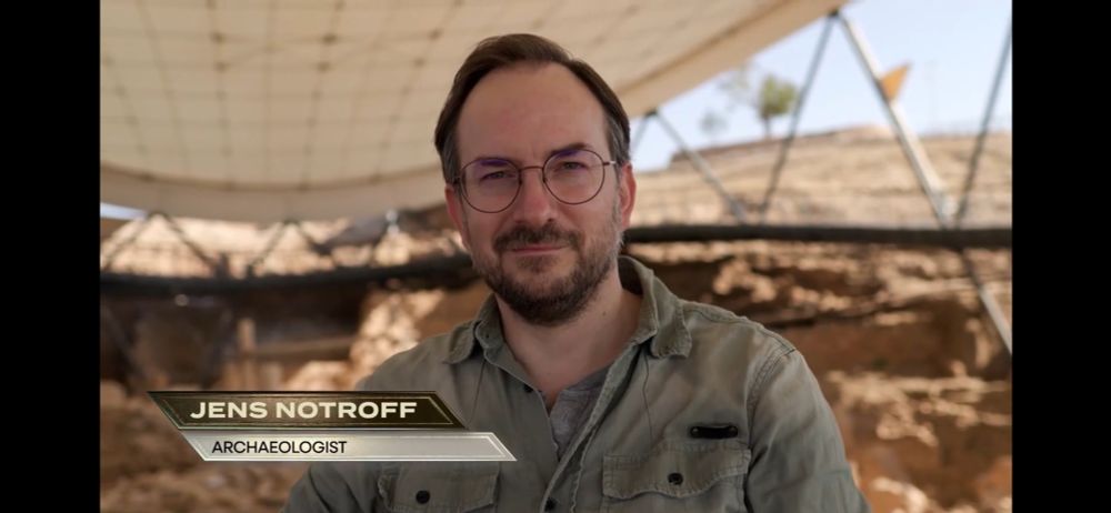 A man with glasses and a beard stands under a canopy in front of an excavation site. On-screen text reads: Jens Notroff, Archaeologist. (Sure, that's me.)