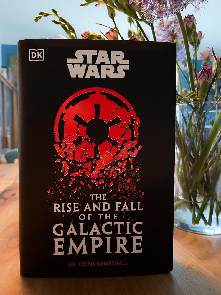 Book cover of Star Wars: The Rise and Fall of the Galactic Empire by Dr Chris Kempshall. The bursting red symbol of the Galactic Empire is depicted on a black background. The title is written in white underneath. Flowers in a vase on the same wooden table as the book can be seen in the background.