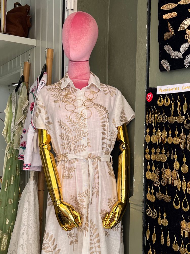 A mannequin with golden arms wears a white dress with gold leaf patterns. 