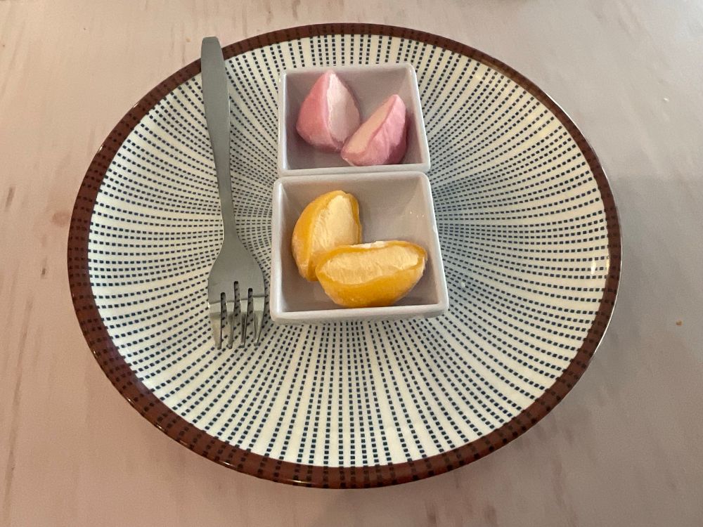 Four halves of mochi on small square plates on top of a bigger round plate. They're orange and strawberry flavoured. There's a fork on the plate and they're on top of a white wooden table.