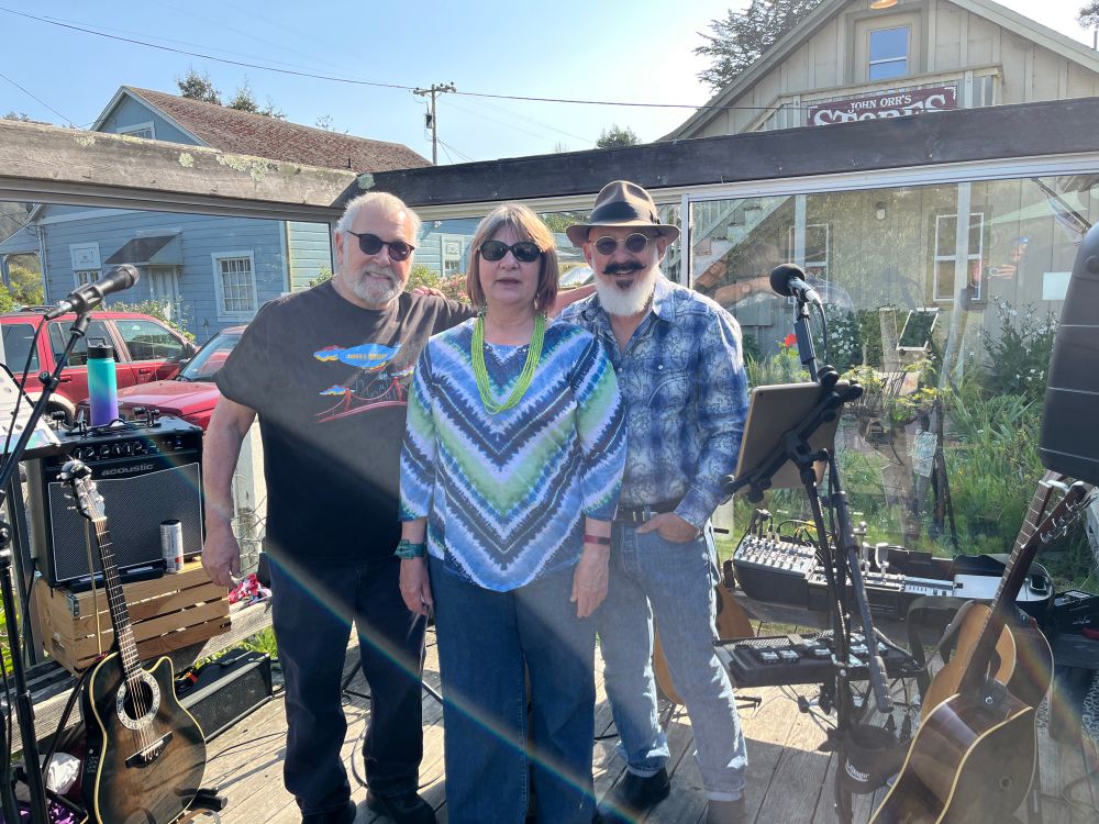 Two standing men and one woman on deck with guitars and mic stands. 