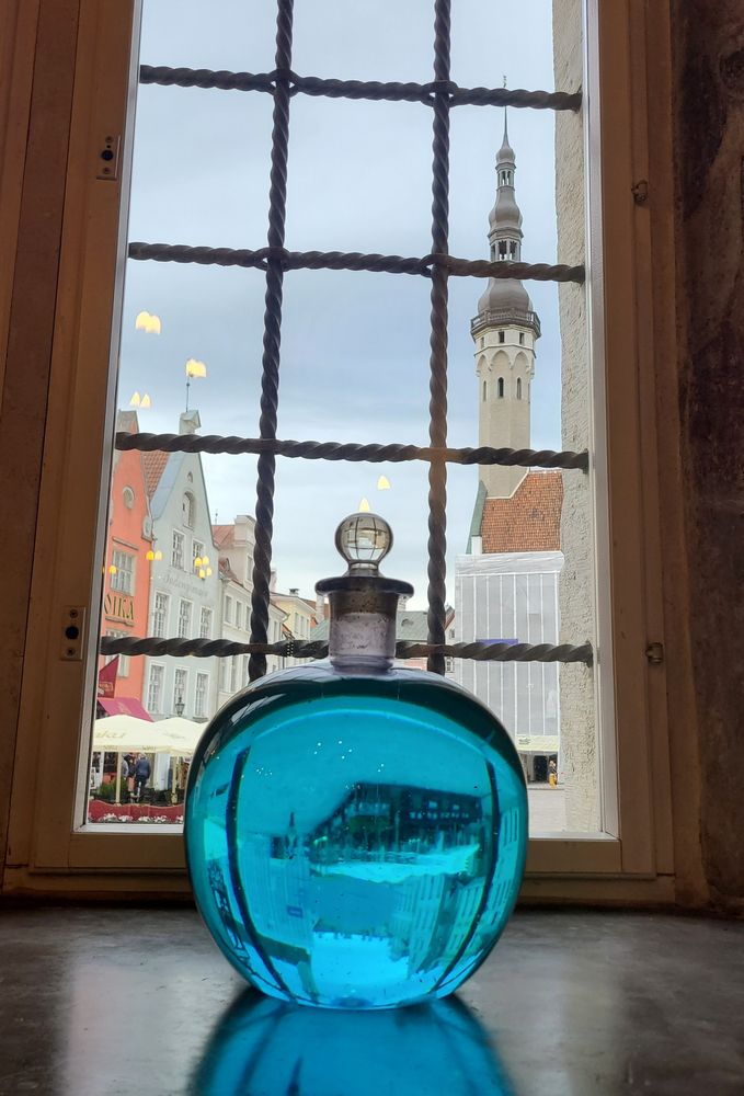 Eine blaue Glaskugel vor einem vergitterten Fenster. Dadurch kann man einen Turm und einige Häusergiebel sehen.