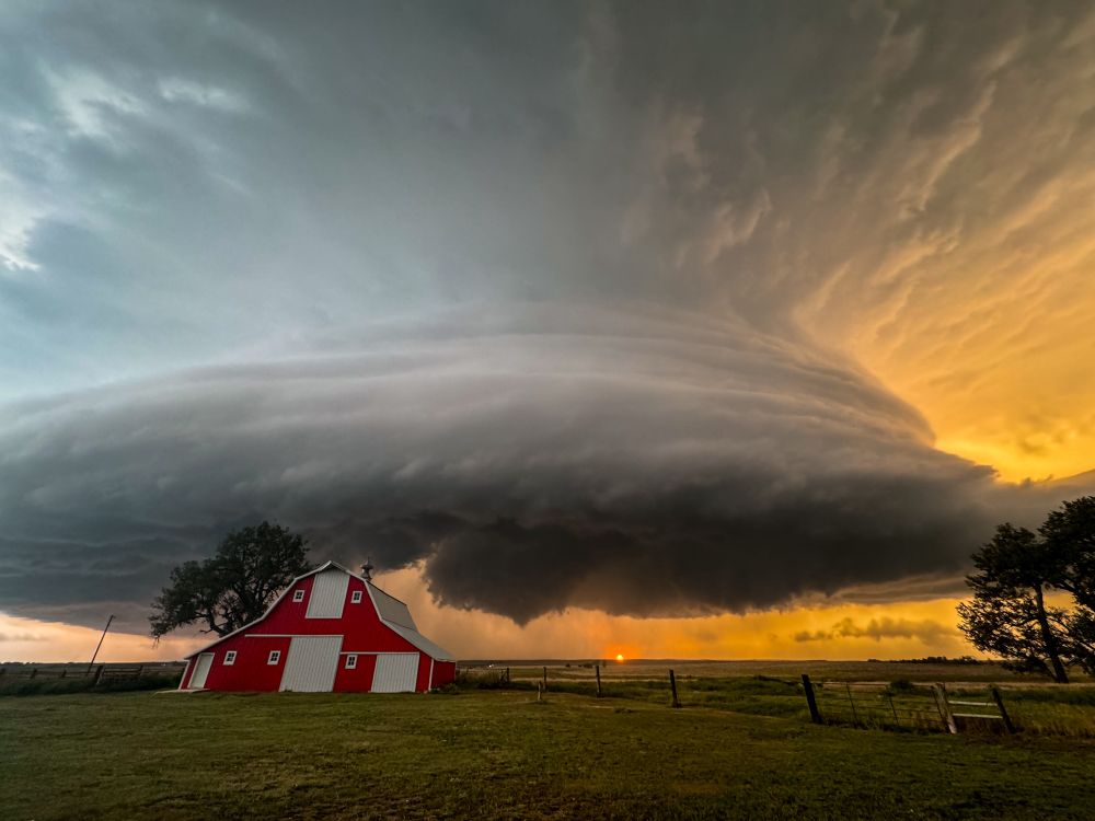 Photo of a supercell thunderstorm with the setting sun below casting a bright orange sky behind the storm and a bright red barn near Mound City, South Dakota on August 28, 2024.