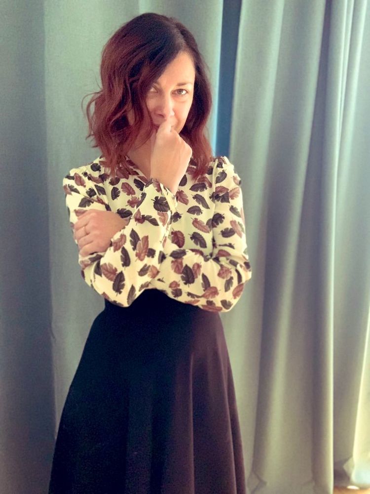 Foto von mir, stehend, ich trage ein Kleid mit einem Blusenteil in beige mit grünen und braunen Blättern und einem schwarzen Rockteil.
Meine Arme sind verschränkt, eine Hand vor dem Mund.