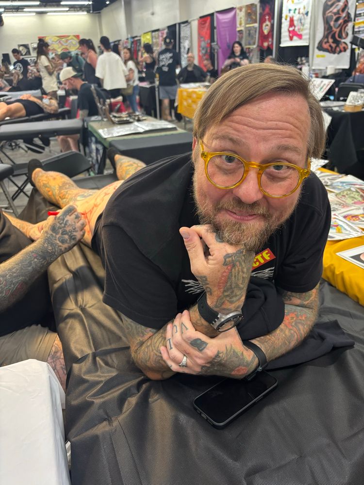 An aging ginger in yellow glasses laying on a table, ass up, being drawn on for a tattoo