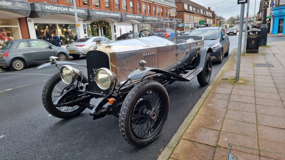 An old car - silver metal, open seating, small windscreen, running boards and narrow wheels. 
