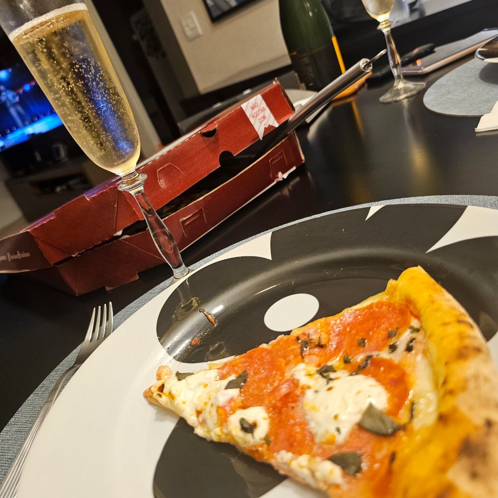 Taça de espumante e fatia de pizza num prato branco e preto. Ao fundo, caixa de pizza vinho, garrafa verde de espumante.